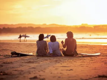 Contiki travellers relaxing on the sand with surfboards watching surfers walk along the shoreline at a stunning golden sunset in Byron Bay Australia
