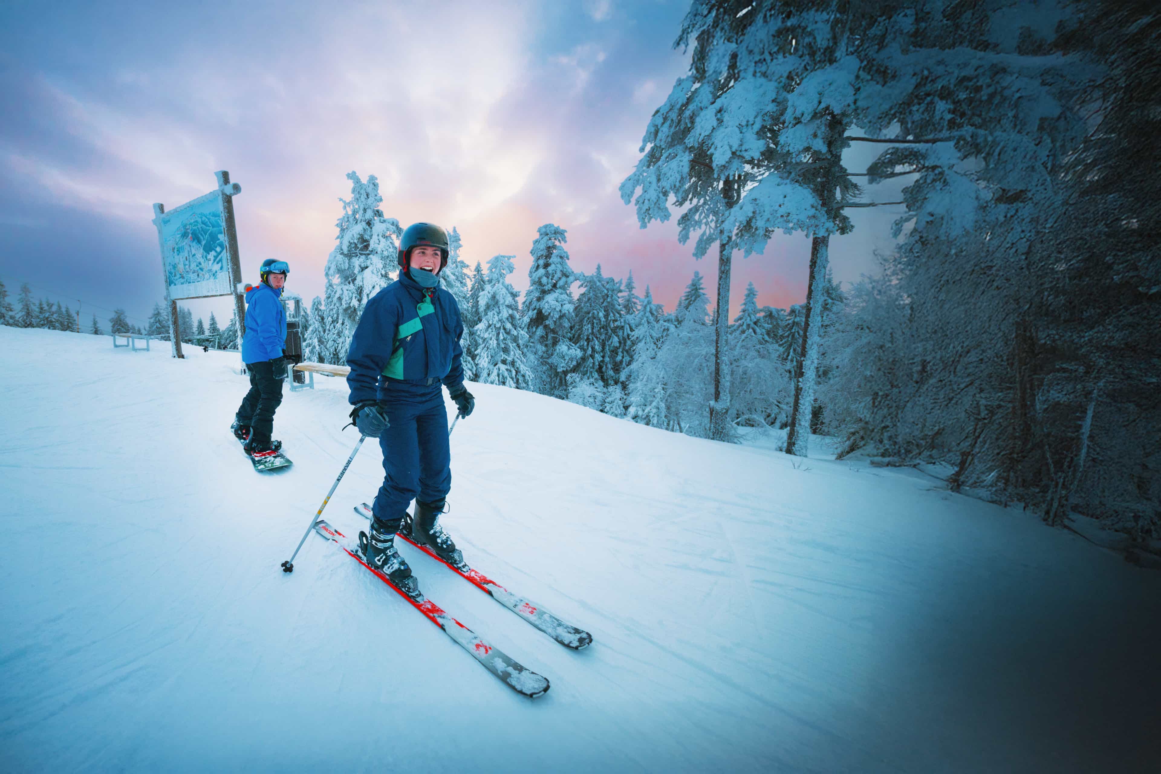 Skier and snowboarder on a snow-covered slope through frosted forest at dusk in Scandinavia.