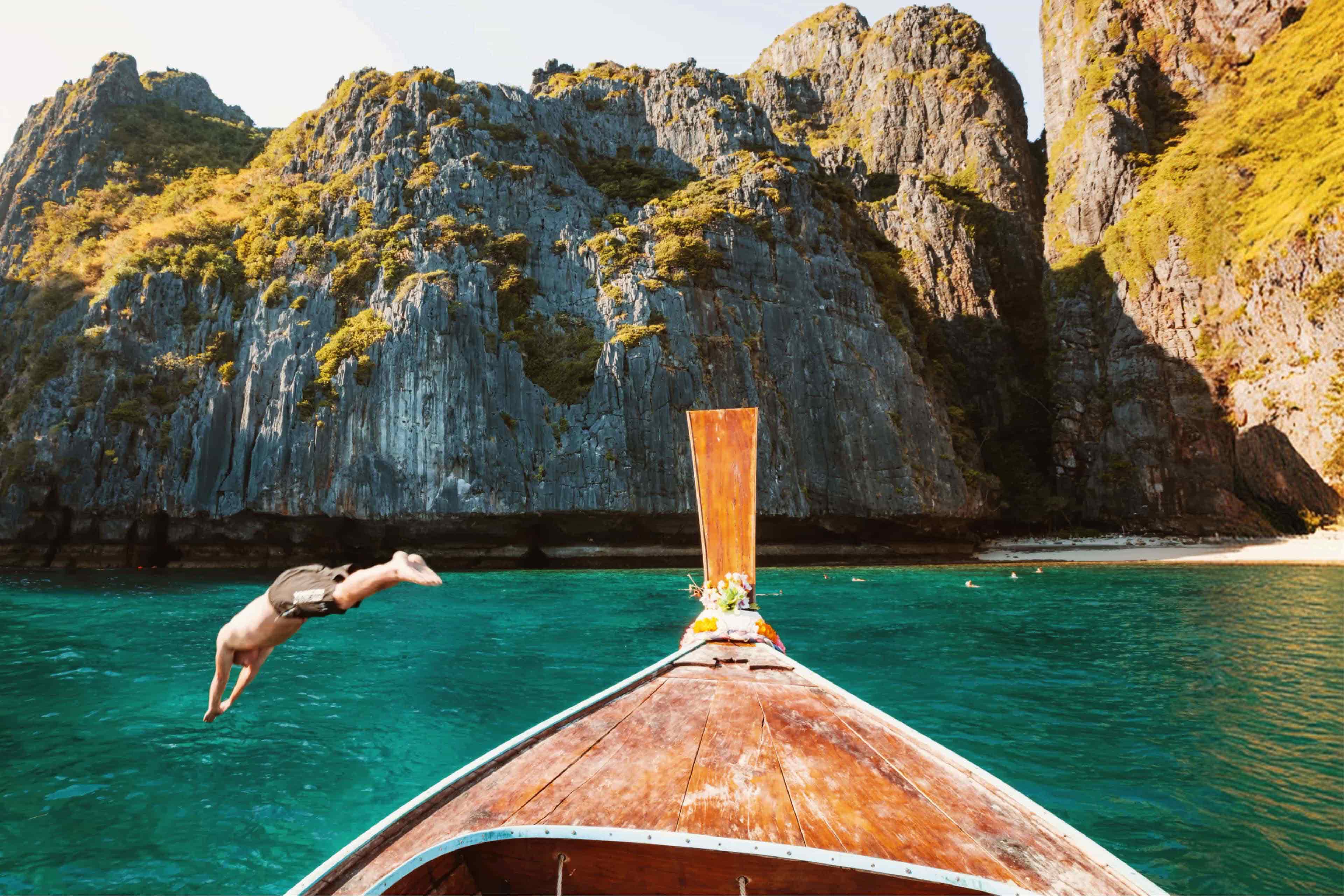 Traveller diving off a longtail boat into turquoise water beneath towering limestone cliffs, Thailand.