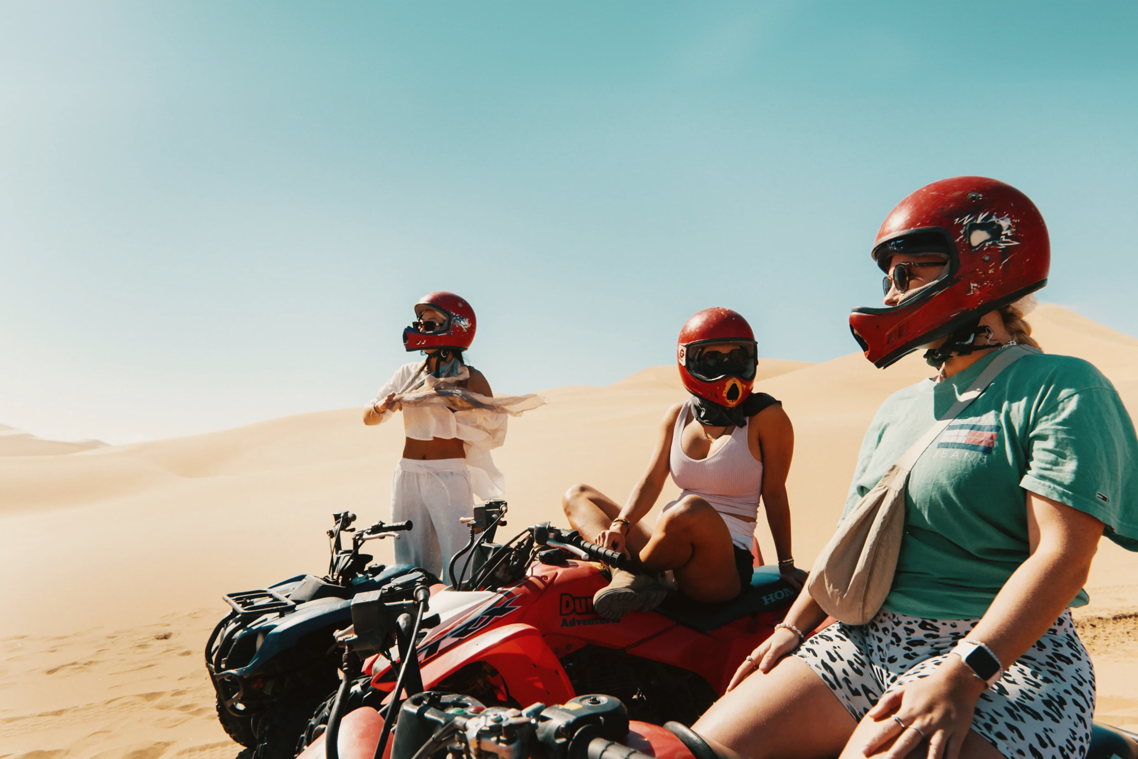Travellers wearing helmets and gathered around quad bikes on sand dunes in the desert.