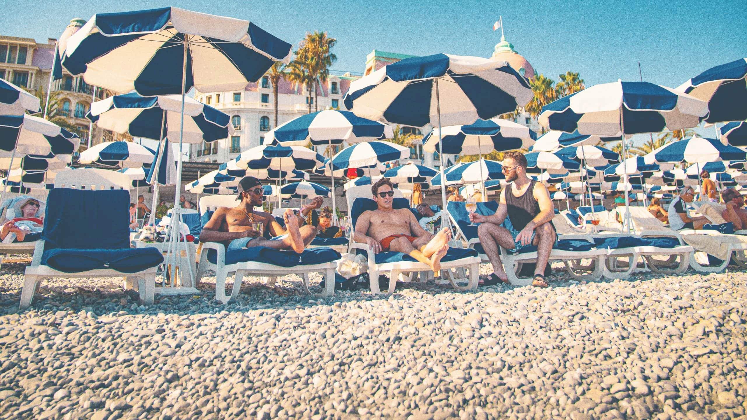 Three travellers unwind on sun loungers beneath striped beach umbrellas along the French Riviera on the Contiki London to Athens with Greek Islands Trip