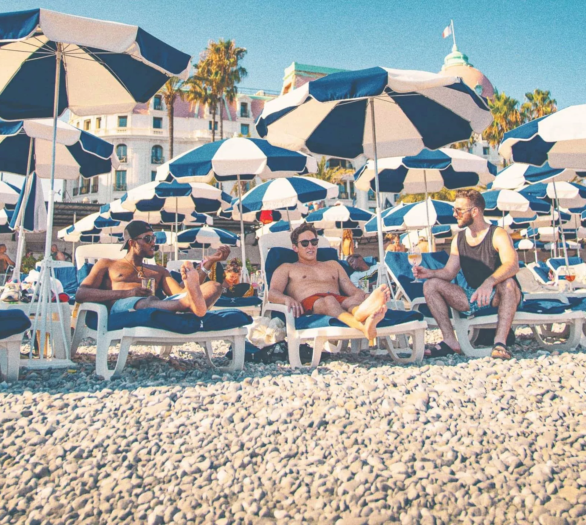 Three travellers unwind on sun loungers beneath striped beach umbrellas along the French Riviera on the Contiki London to Athens with Greek Islands Trip