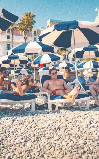Three travellers unwind on sun loungers beneath striped beach umbrellas along the French Riviera on the Contiki London to Athens with Greek Islands Trip