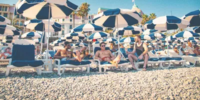 Three travellers unwind on sun loungers beneath striped beach umbrellas along the French Riviera on the Contiki London to Athens with Greek Islands Trip