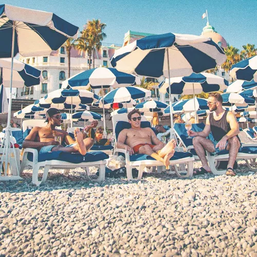 Three travellers unwind on sun loungers beneath striped beach umbrellas along the French Riviera on the Contiki London to Athens with Greek Islands Trip