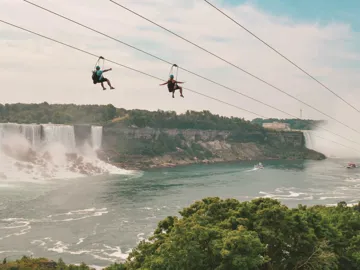 Two travellers ziplining above Niagara Falls gorge with the American and Horseshoe Falls and misty river below - Contiki Best of Canada Tour