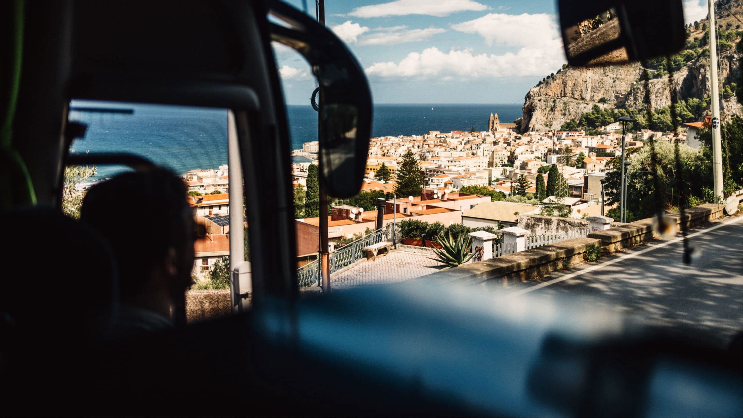 View from a coach overlooking the coastal town of Cefalù in Sicily, with terracotta rooftops, dramatic cliffs and the Mediterranean Sea beyond - Sicily Escape Tour