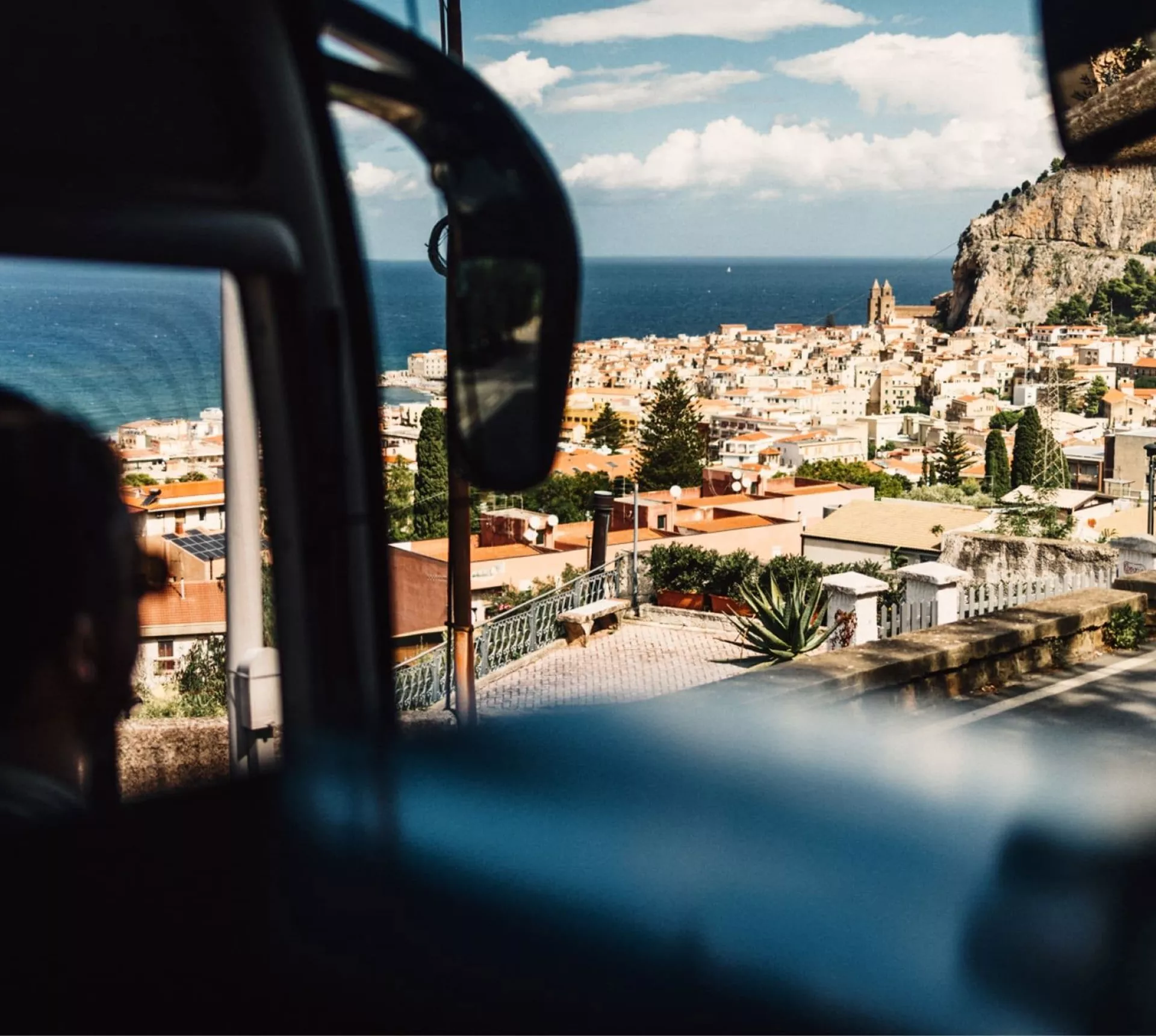 View from a coach overlooking the coastal town of Cefalù in Sicily, with terracotta rooftops, dramatic cliffs and the Mediterranean Sea beyond - Sicily Escape Tour