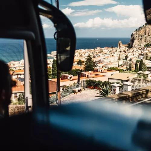 View from a coach overlooking the coastal town of Cefalù in Sicily, with terracotta rooftops, dramatic cliffs and the Mediterranean Sea beyond - Sicily Escape Tour