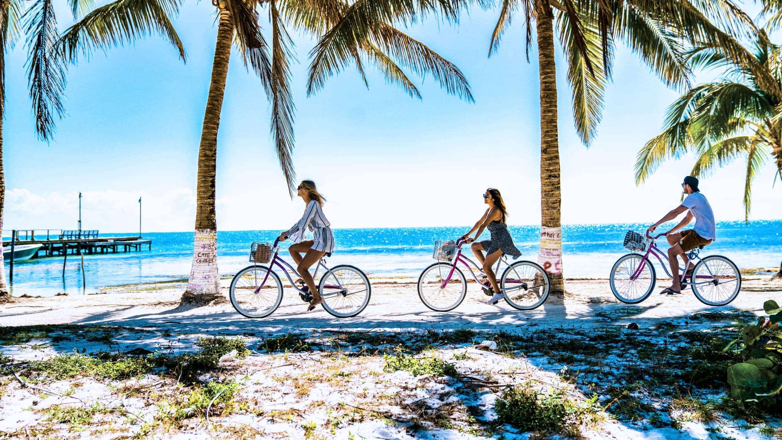 Three young travellers cycling on beach cruiser bikes along the sandy palm-lined seafront of Caye Caulker island in Belize - Contiki Mexico City to San Jose Tour