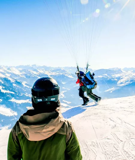 Two skiers in helmets watching a paraglider launch from a snowy Alpine summit with breathtaking panoramic views of the Austrian Alps mountain range - Contiki Ski Austria Tour