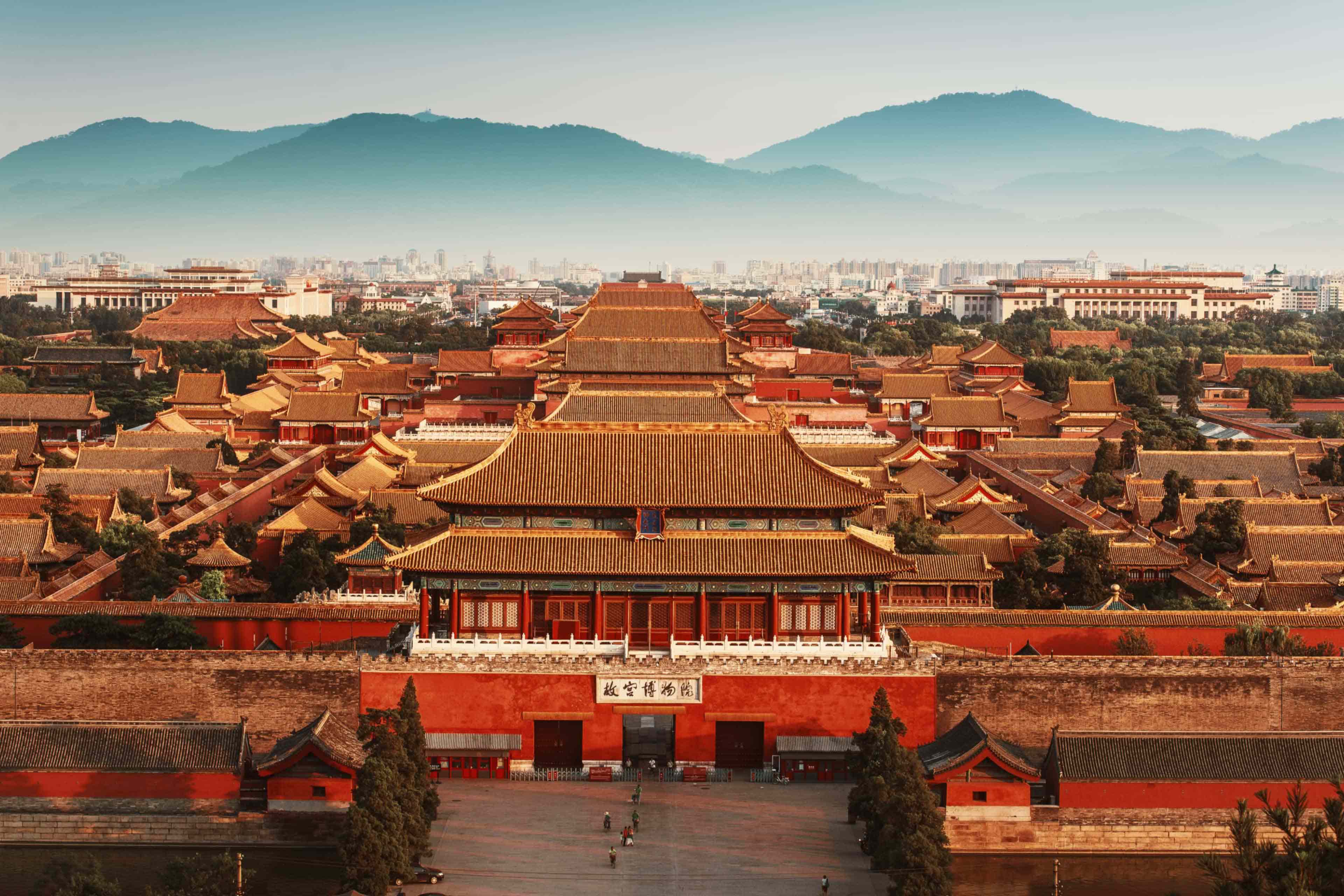Aerial view of the Forbidden City's golden-roofed palace complex in Beijing, with the modern city skyline and mountains beyond.