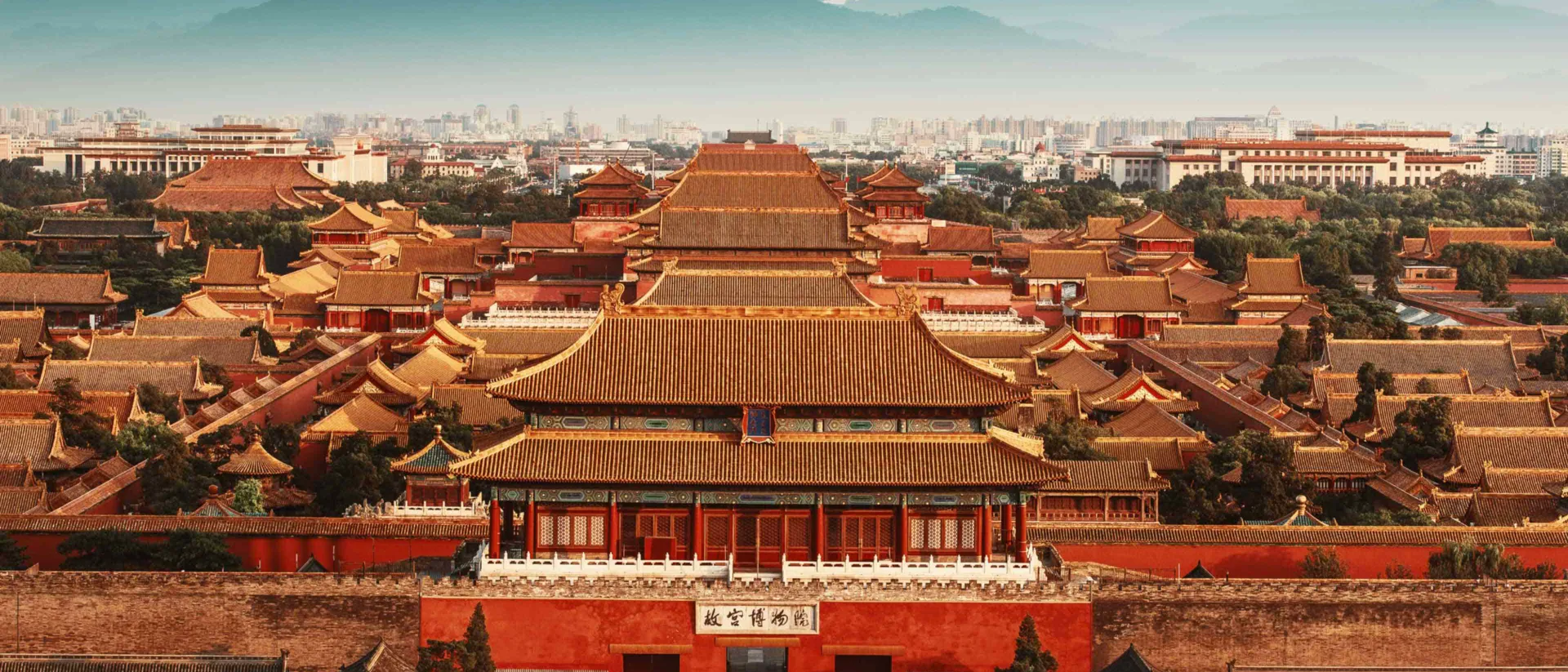 Aerial view of the Forbidden City's golden-roofed palace complex in Beijing, with the modern city skyline and mountains beyond.