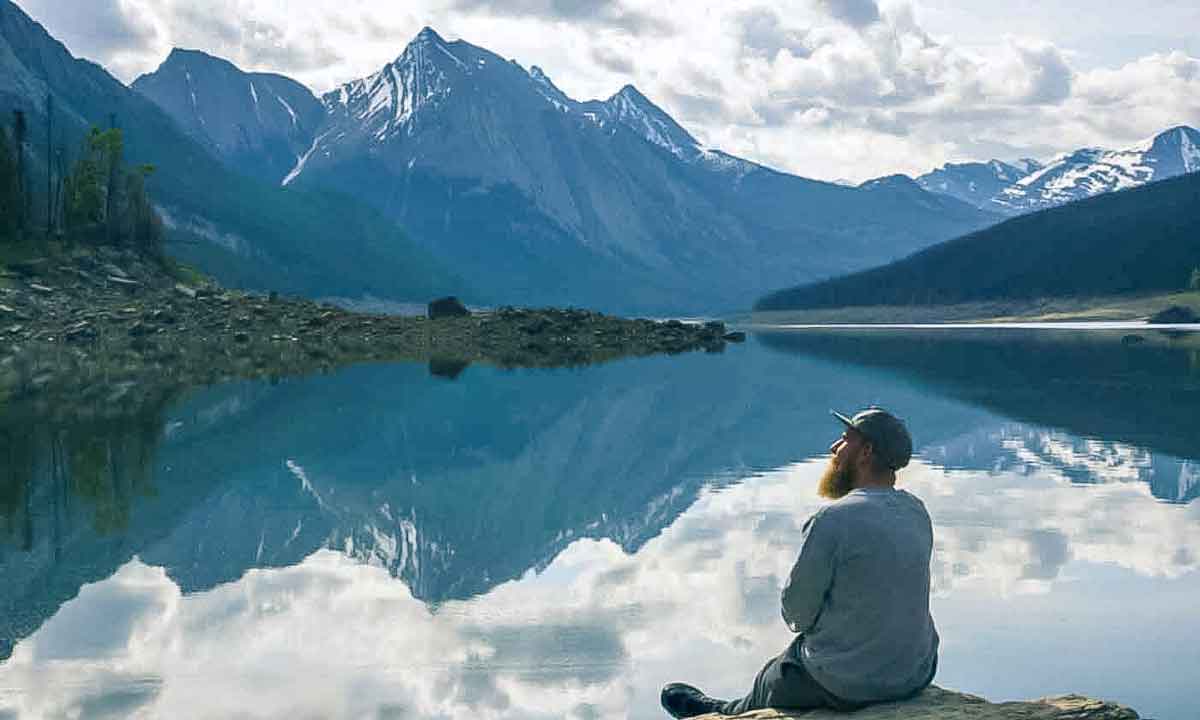 Man Sitting Close O A Lake Looking At Snowy Mountains