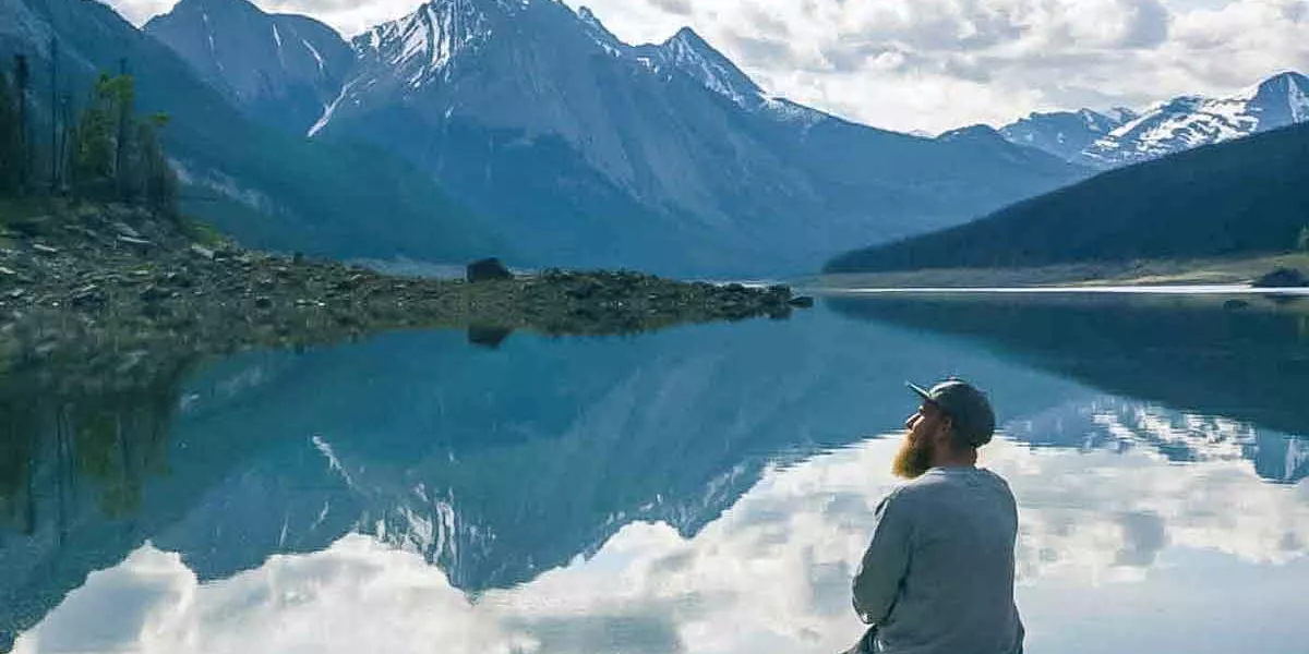 Man Sitting Close O A Lake Looking At Snowy Mountains