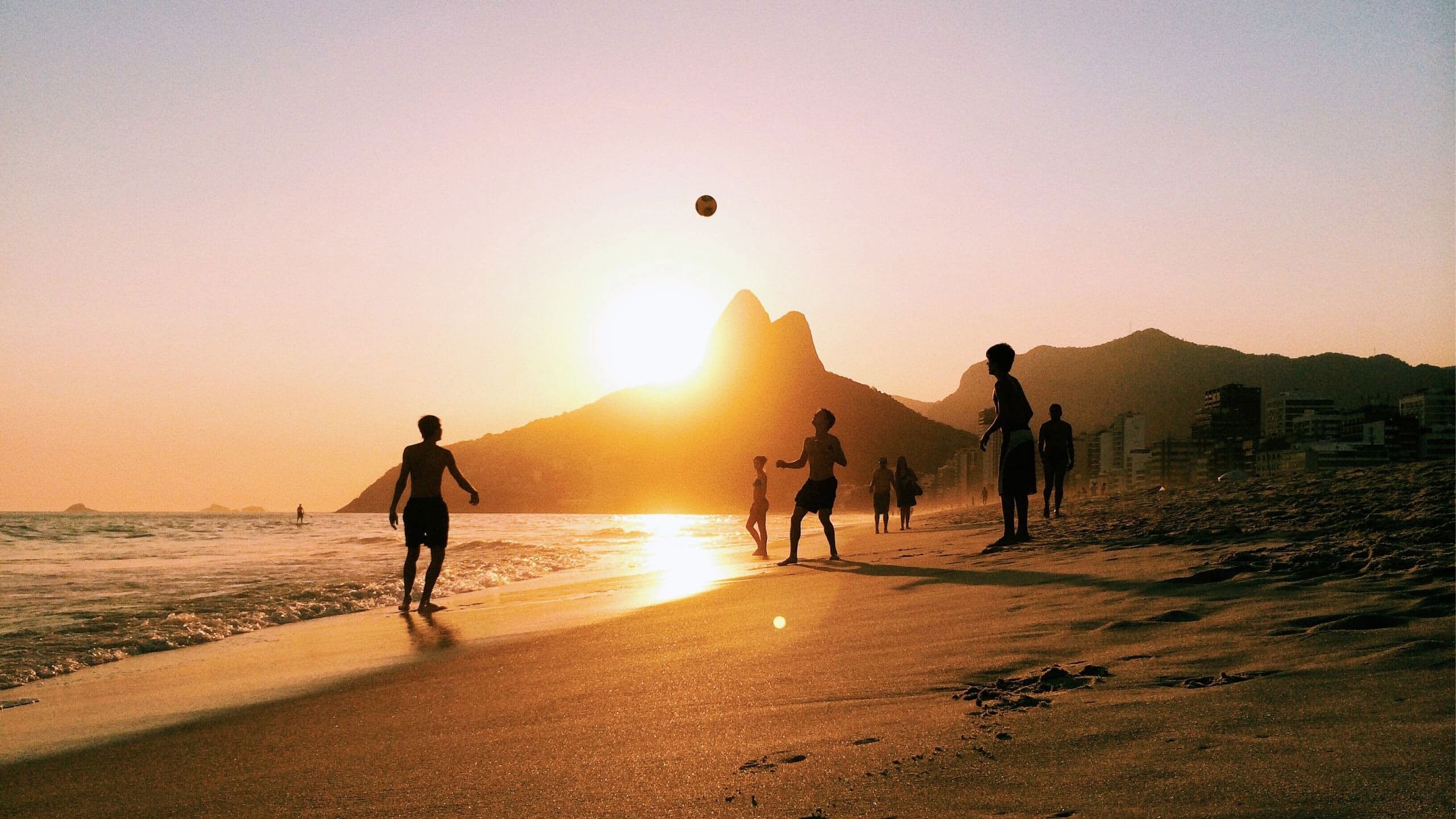 People enjoy a game of beach volleyball as the sun sets over Rio de Janeiro, with golden light reflecting on the sand and the city's dramatic mountain skyline beyond