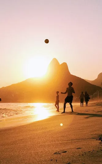 People enjoy a game of beach volleyball as the sun sets over Rio de Janeiro, with golden light reflecting on the sand and the city's dramatic mountain skyline beyond