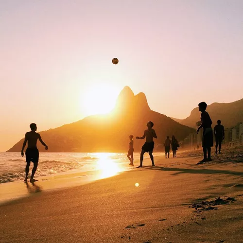 People enjoy a game of beach volleyball as the sun sets over Rio de Janeiro, with golden light reflecting on the sand and the city's dramatic mountain skyline beyond