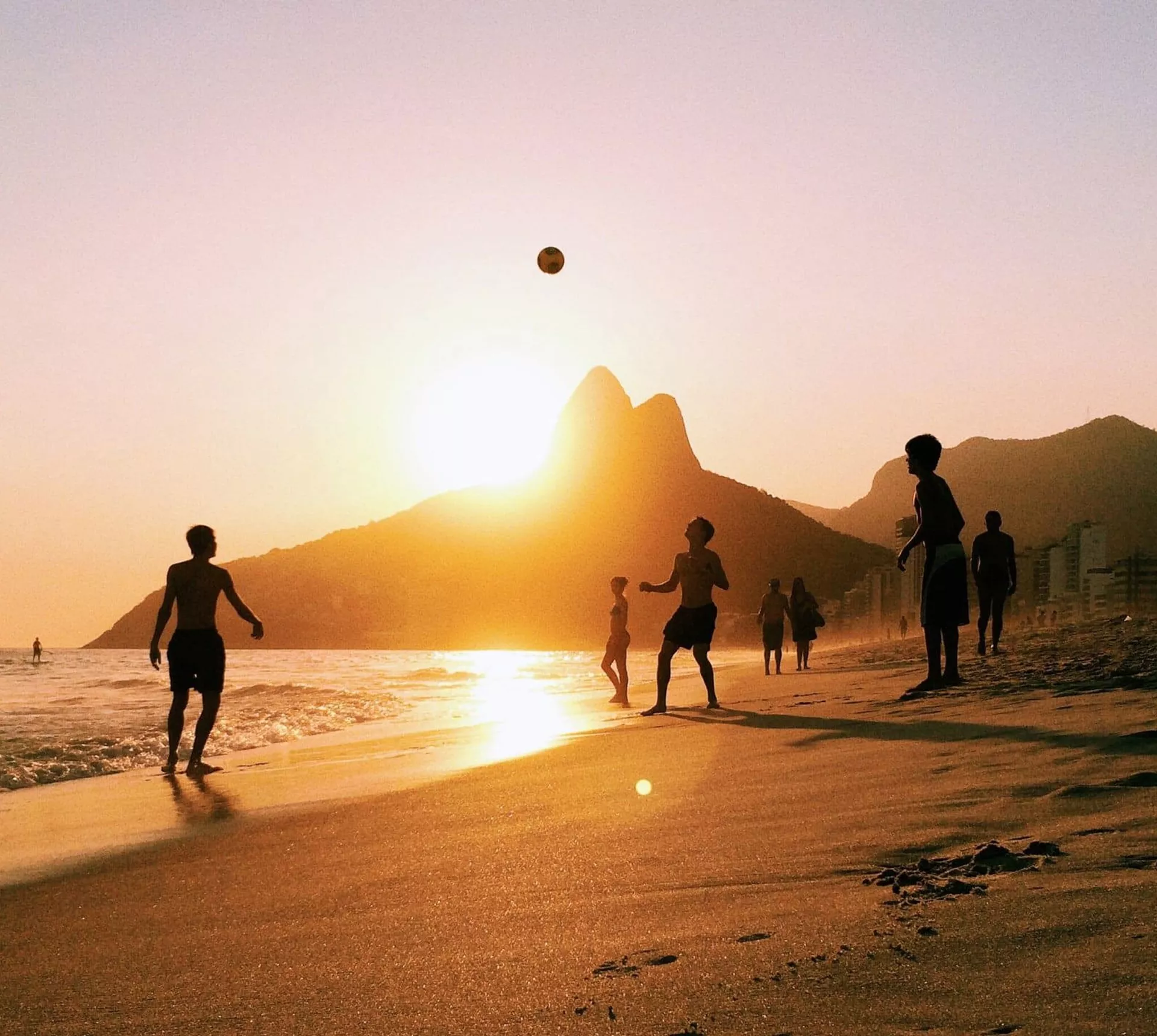 People enjoy a game of beach volleyball as the sun sets over Rio de Janeiro, with golden light reflecting on the sand and the city's dramatic mountain skyline beyond
