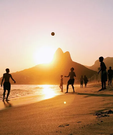 People enjoy a game of beach volleyball as the sun sets over Rio de Janeiro, with golden light reflecting on the sand and the city's dramatic mountain skyline beyond