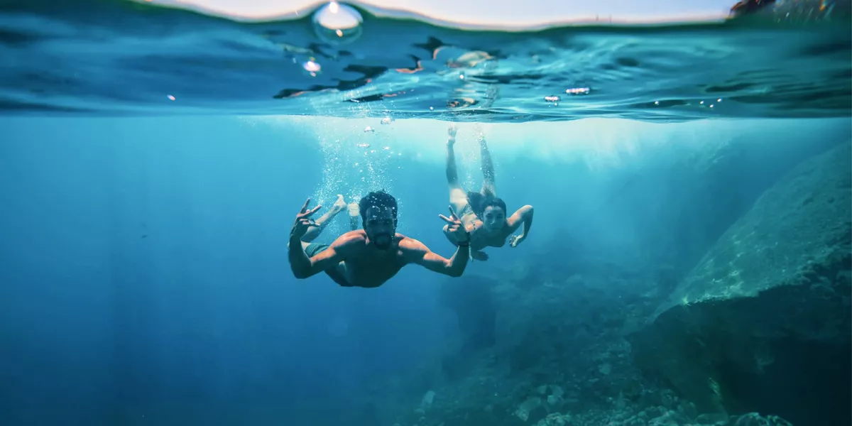 Two young travellers freediving underwater making peace signs in the clear blue waters of the Greek Islands - Contiki Athens and Santorini island hopper Tour