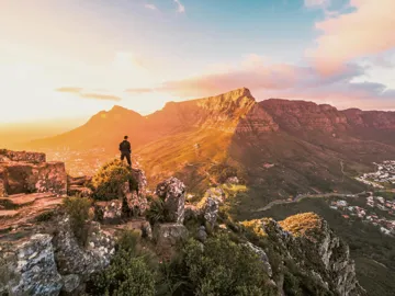 Solo traveller standing on rocky Lion's Head summit at golden hour overlooking the iconic Table Mountain and Cape Town cityscape in South Africa - Contiki Cape Town, Safari & Victoria Falls Tour