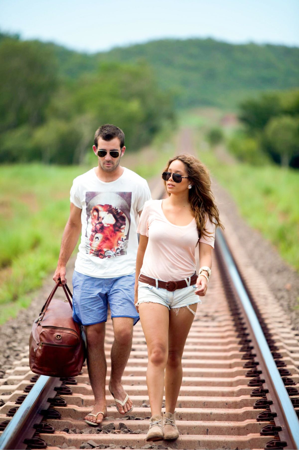 Couple wearing summer clothes and sunglasses walking on train tracks through green land
