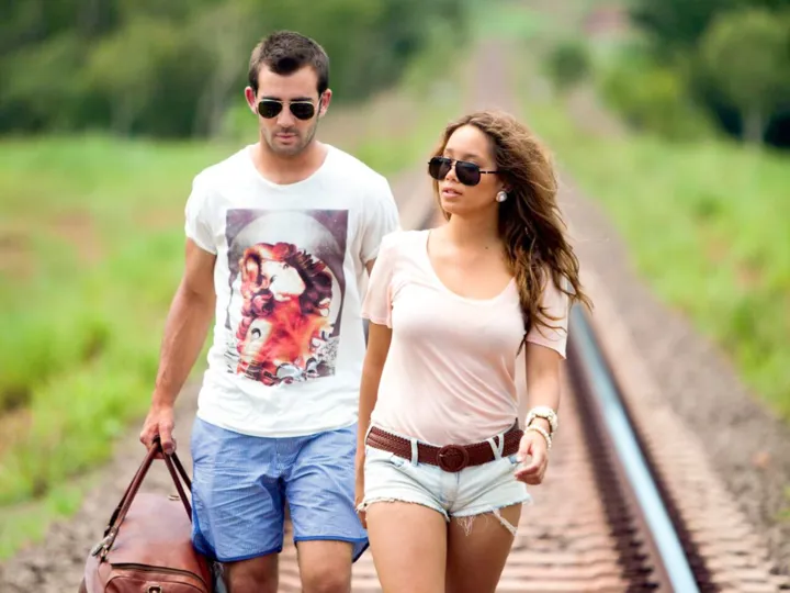 Couple wearing summer clothes and sunglasses walking on train tracks through green land