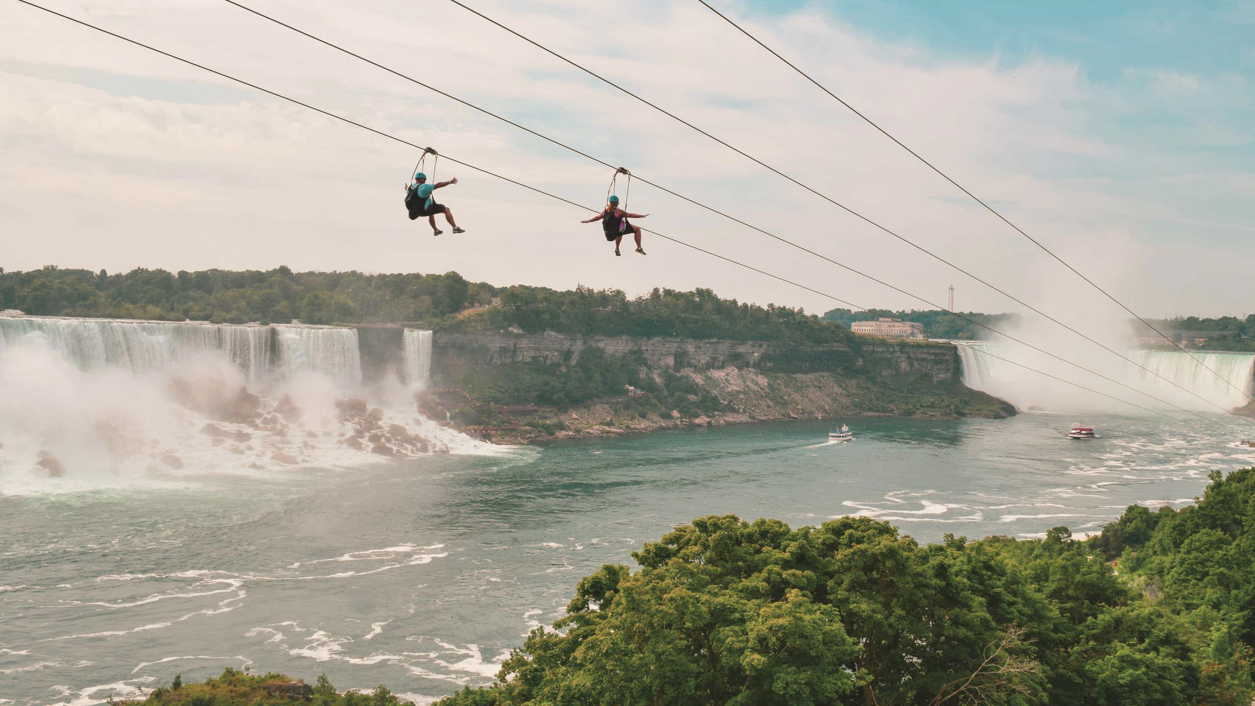 Two travellers ziplining above Niagara Falls gorge with the American and Horseshoe Falls and misty river below - Contiki Best of Canada Tour