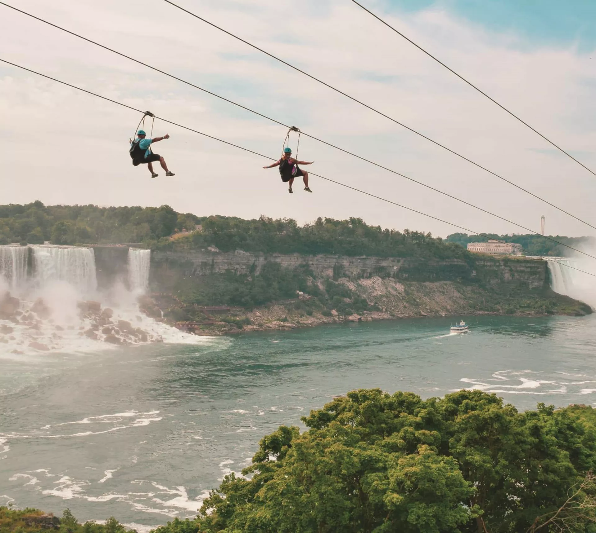 Two travellers ziplining above Niagara Falls gorge with the American and Horseshoe Falls and misty river below - Contiki Best of Canada Tour