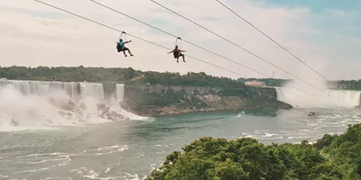 Two travellers ziplining above Niagara Falls gorge with the American and Horseshoe Falls and misty river below - Contiki Best of Canada Tour