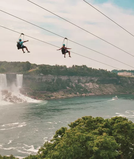 Two travellers ziplining above Niagara Falls gorge with the American and Horseshoe Falls and misty river below - Contiki Best of Canada Tour