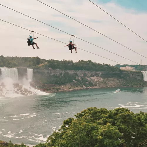 Two travellers ziplining above Niagara Falls gorge with the American and Horseshoe Falls and misty river below - Contiki Best of Canada Tour