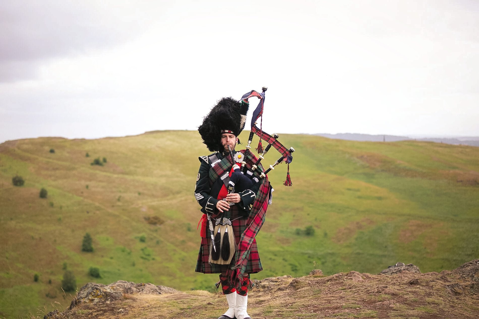 A musician playing bagpipes with green mountains as a background in Scotland.