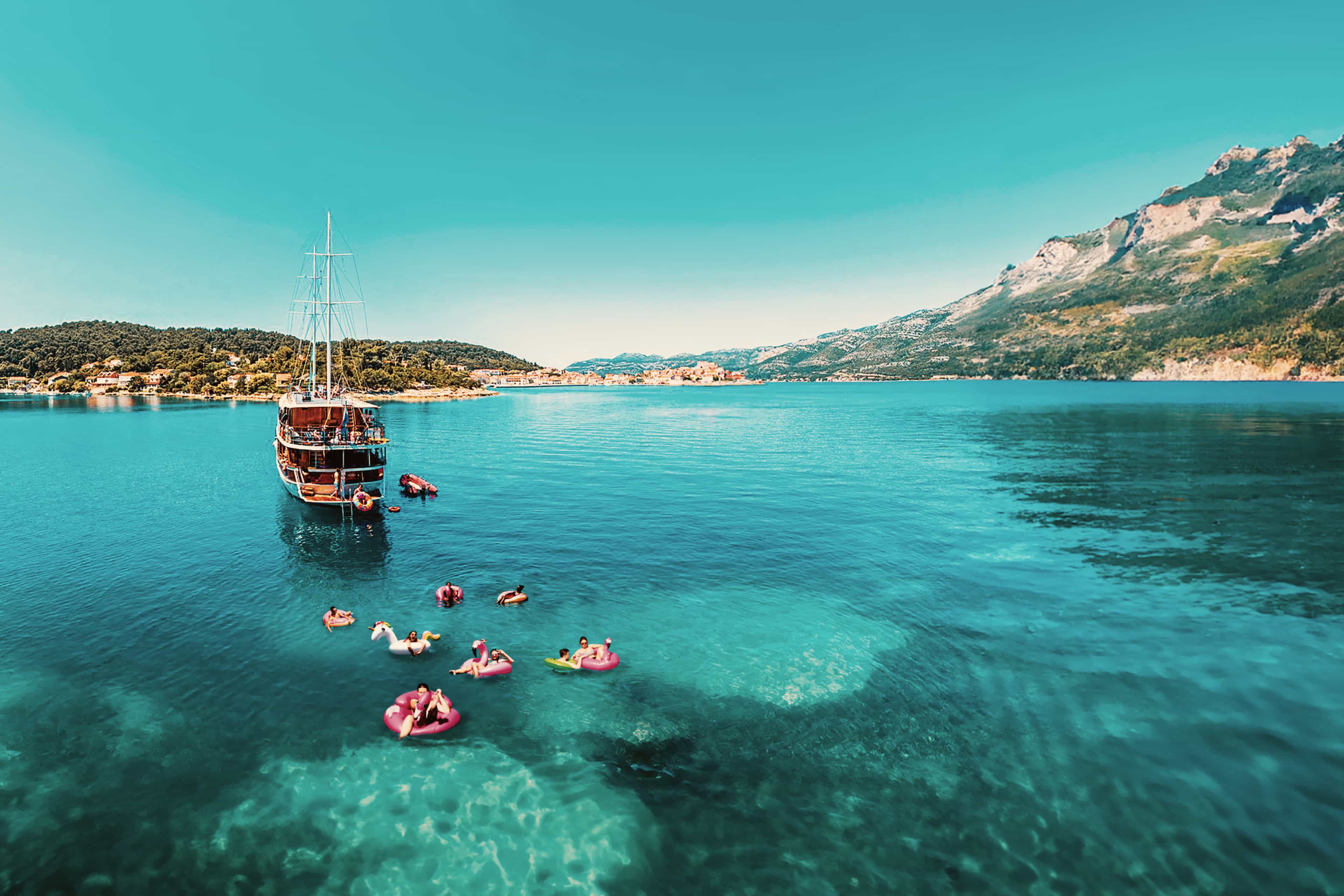 Travellers relaxing on inflatable floats beside a sailing boat in a turquoise bay along the Croatian coast.