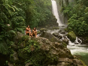 Two travellers relax on rocks beside a rainforest waterfall in Costa Rica, surrounded by lush jungle and a flowing river - Contiki Best of Costa Rica Tour