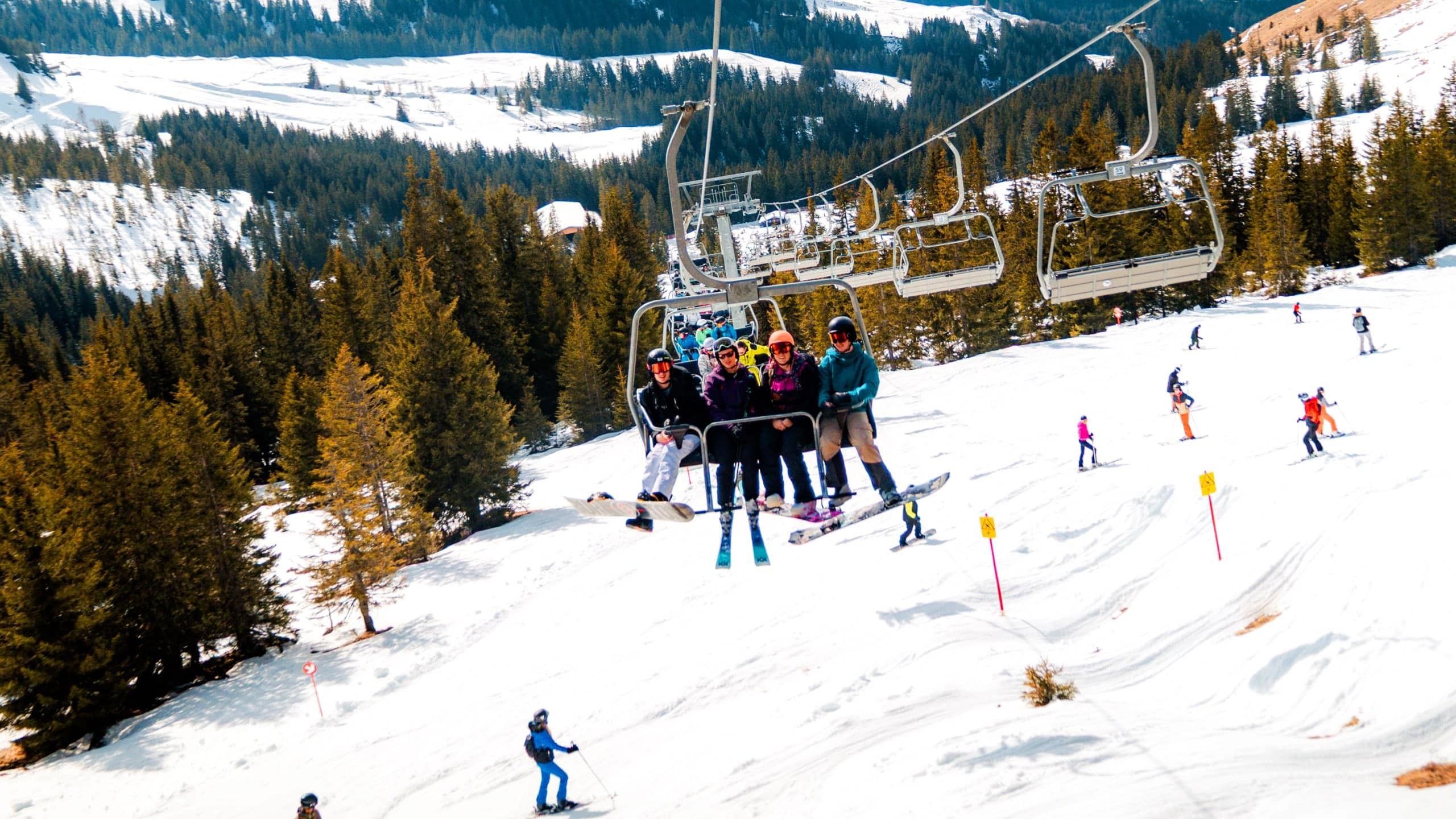 A group of skiers and snowboarders ride a chairlift above snow-covered ski slopes in the Austrian Alps, with pine forests, rolling mountains and other skiers enjoying the runs below.