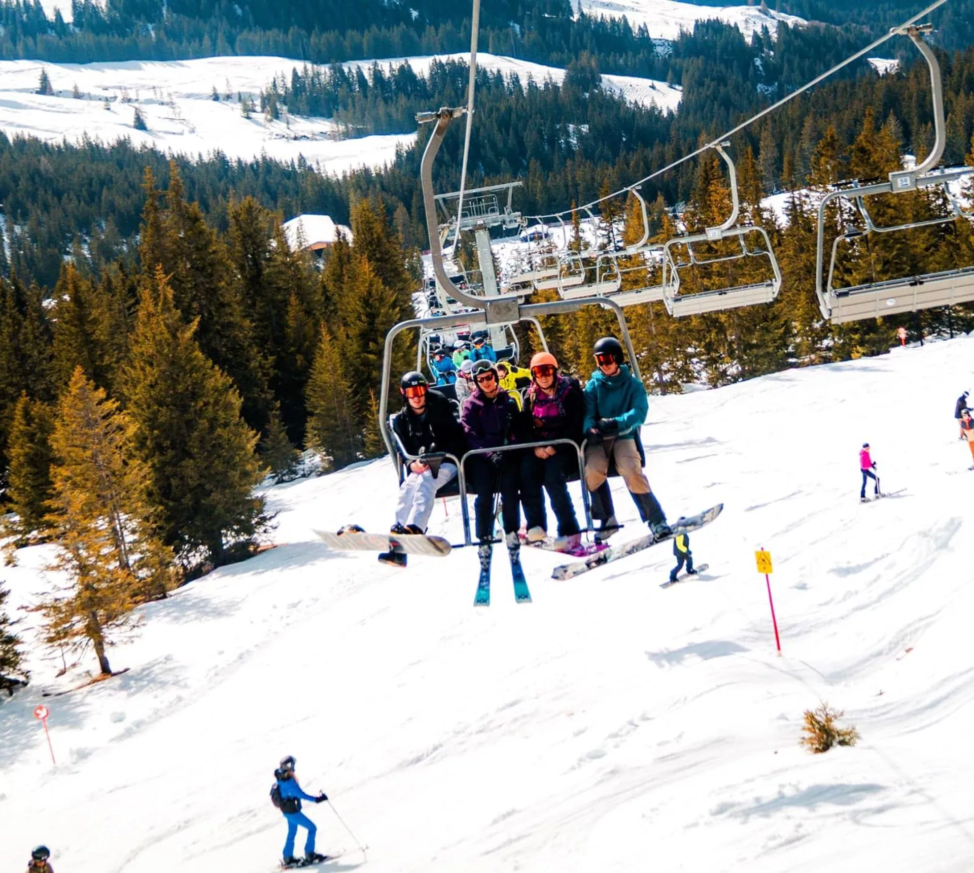 A group of skiers and snowboarders ride a chairlift above snow-covered ski slopes in the Austrian Alps, with pine forests, rolling mountains and other skiers enjoying the runs below.