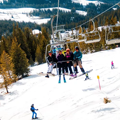 A group of skiers and snowboarders ride a chairlift above snow-covered ski slopes in the Austrian Alps, with pine forests, rolling mountains and other skiers enjoying the runs below.