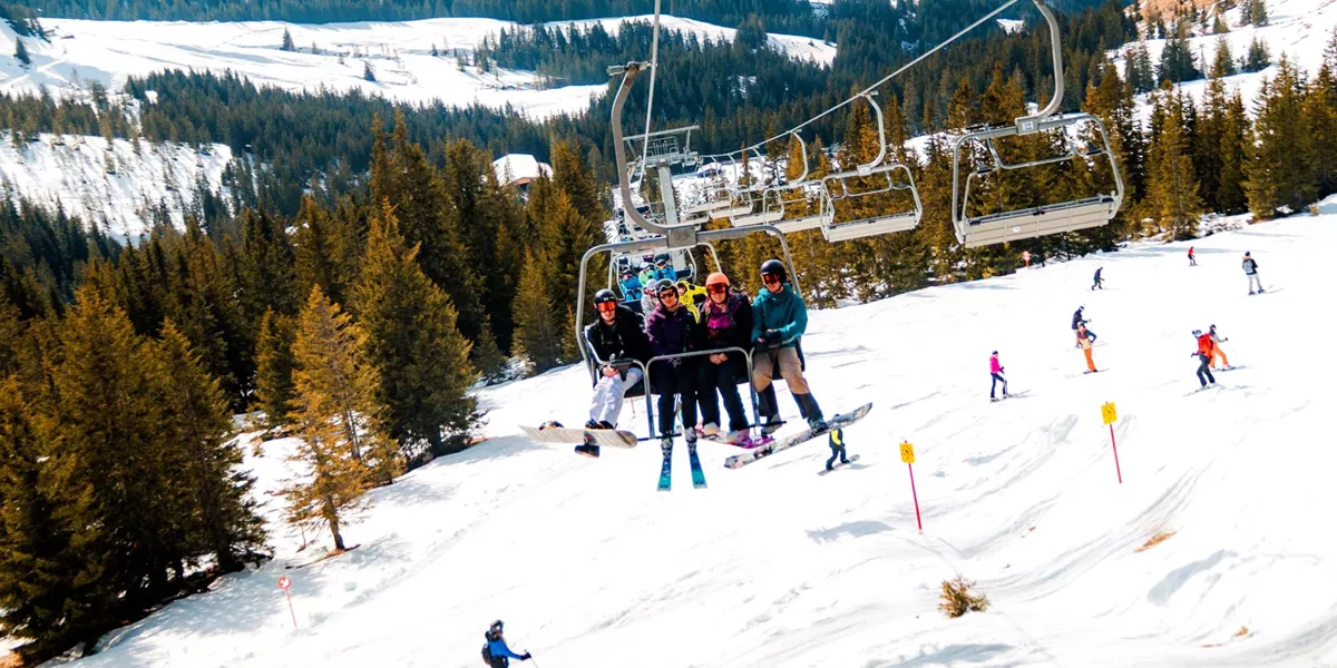 A group of skiers and snowboarders ride a chairlift above snow-covered ski slopes in the Austrian Alps, with pine forests, rolling mountains and other skiers enjoying the runs below.