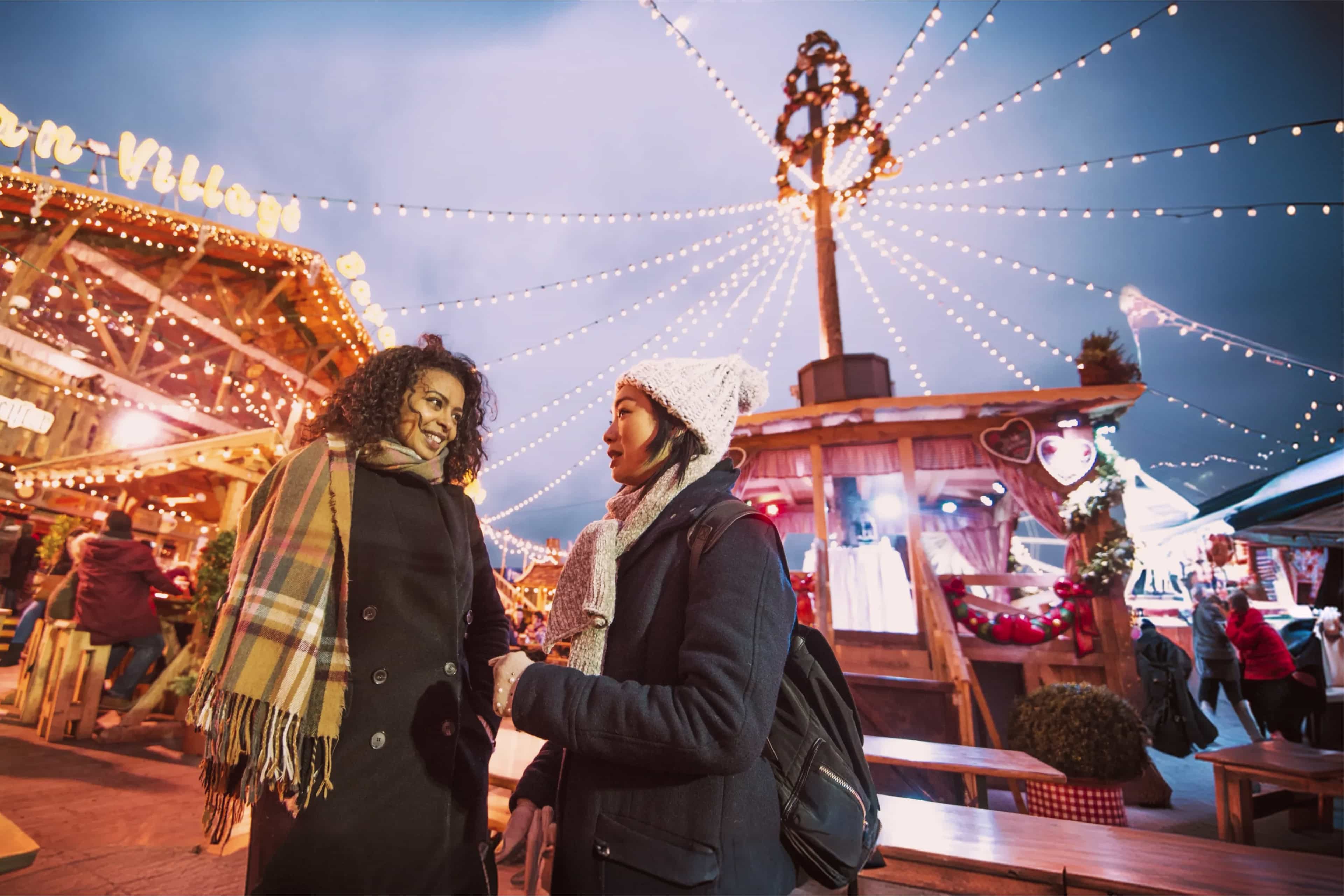 Two travellers wrapped up warm chatting at a festive winter market lit with fairy lights, United Kingdom.