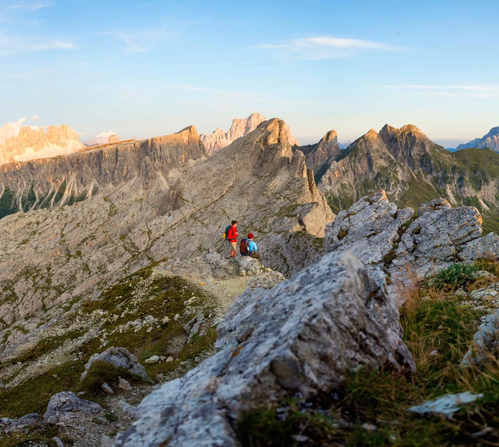 Two hikers with backpacks on a rocky mountain ridge trail at sunset with the dramatic jagged limestone peaks of the Dolomites stretching across the horizon in northern Italy - Contiki Hiking the Dolomites Tour