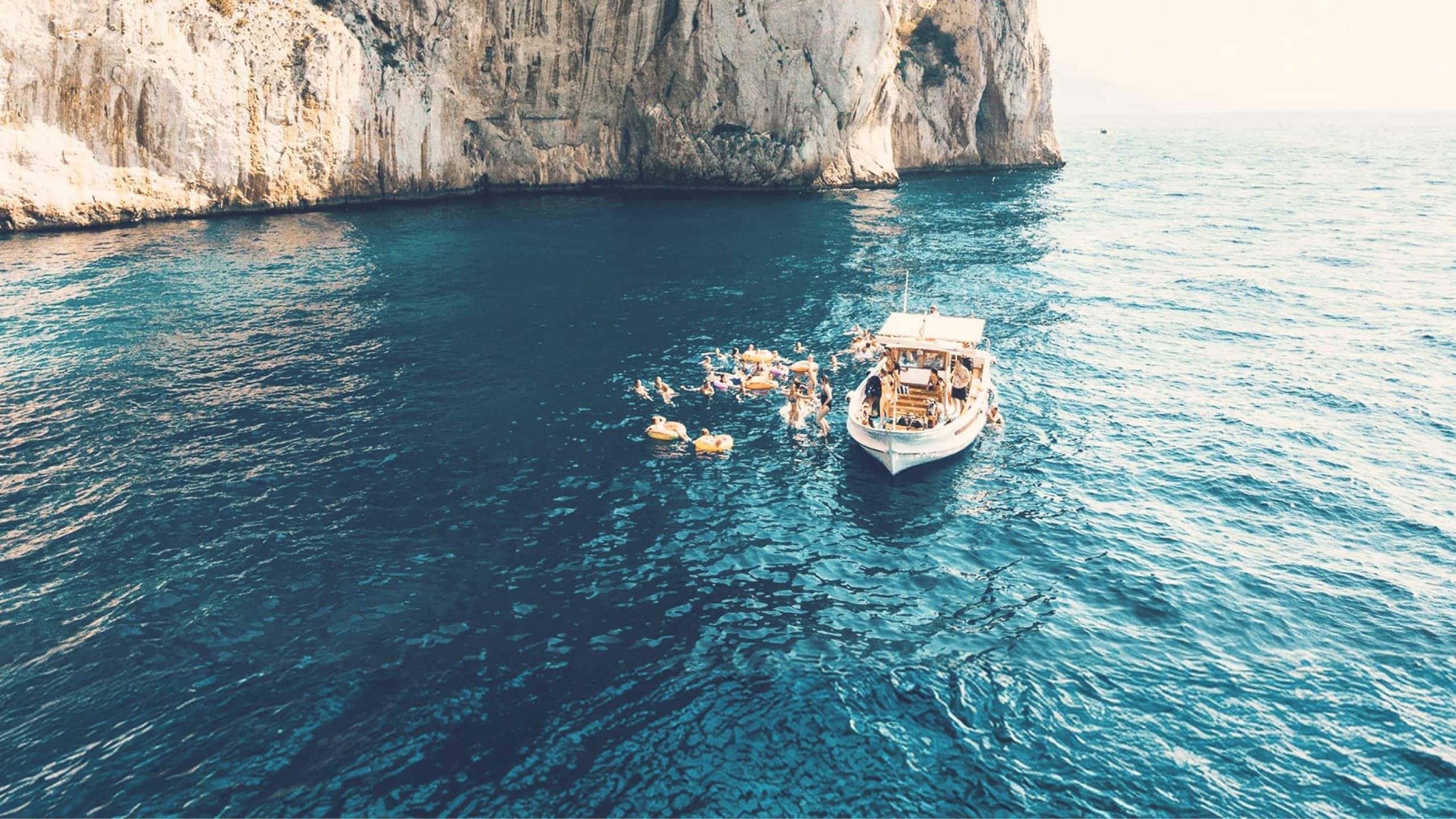 Contiki travellers swimming from a boat beneath the towering cliffs of Capri, Italy, on the Ultimate European Plus Greek Islands Tour