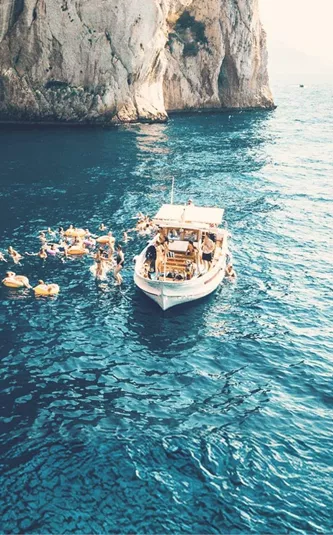 Contiki travellers swimming from a boat beneath the towering cliffs of Capri, Italy, on the Ultimate European Plus Greek Islands Tour