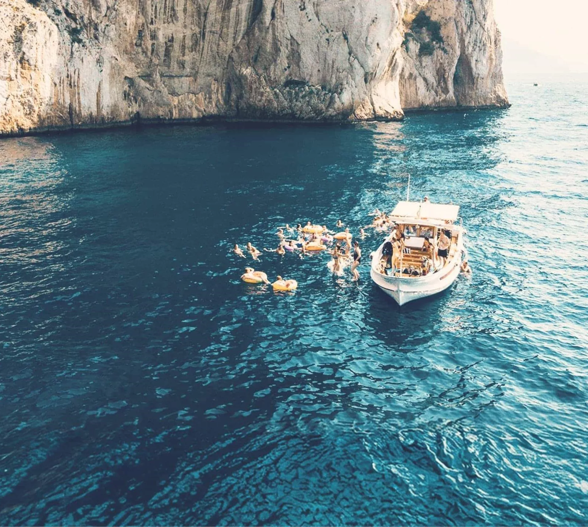 Contiki travellers swimming from a boat beneath the towering cliffs of Capri, Italy, on the Ultimate European Plus Greek Islands Tour