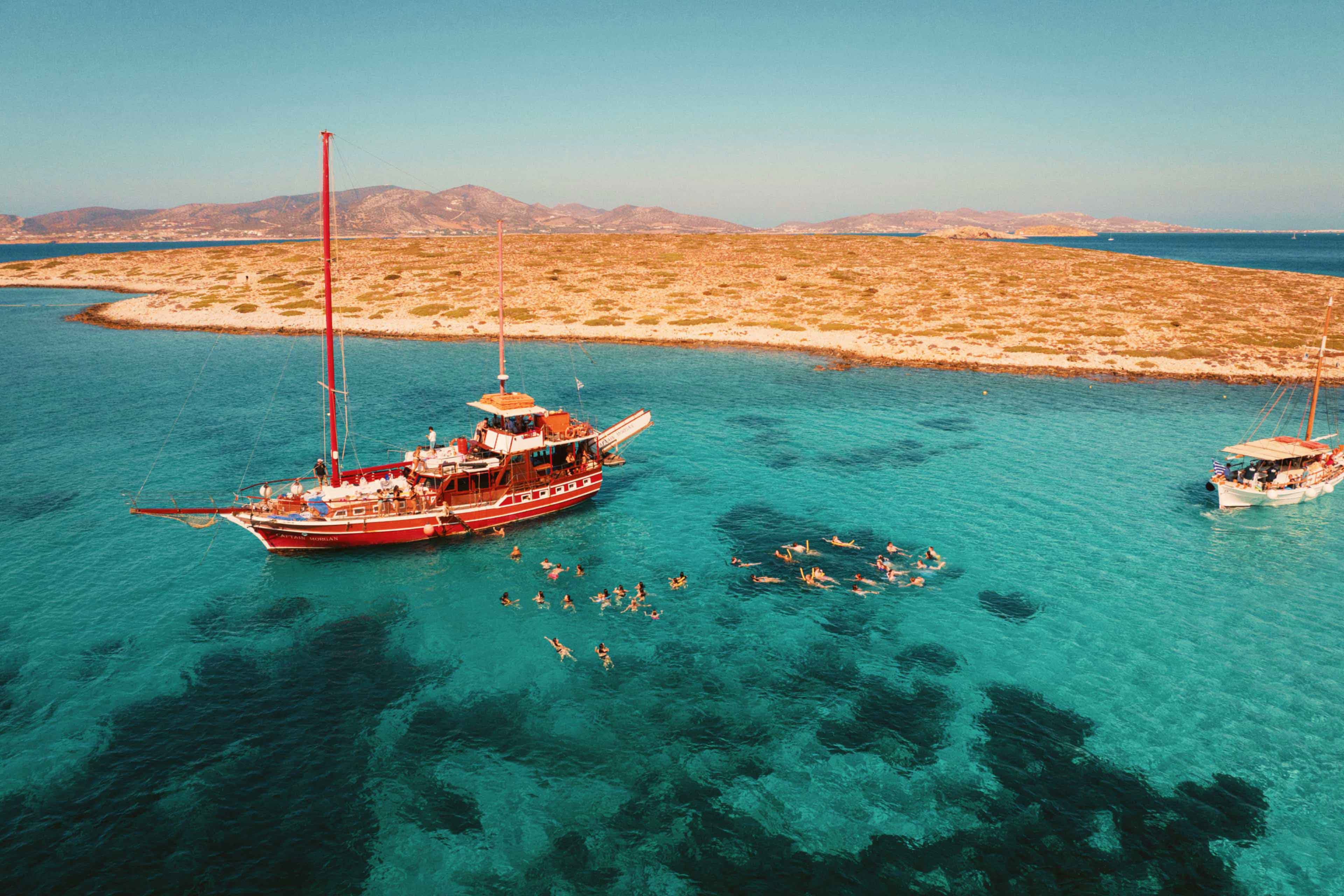 Aerial view of travellers swimming beside a traditional sailing boat in clear turquoise water off the Greek islands.
