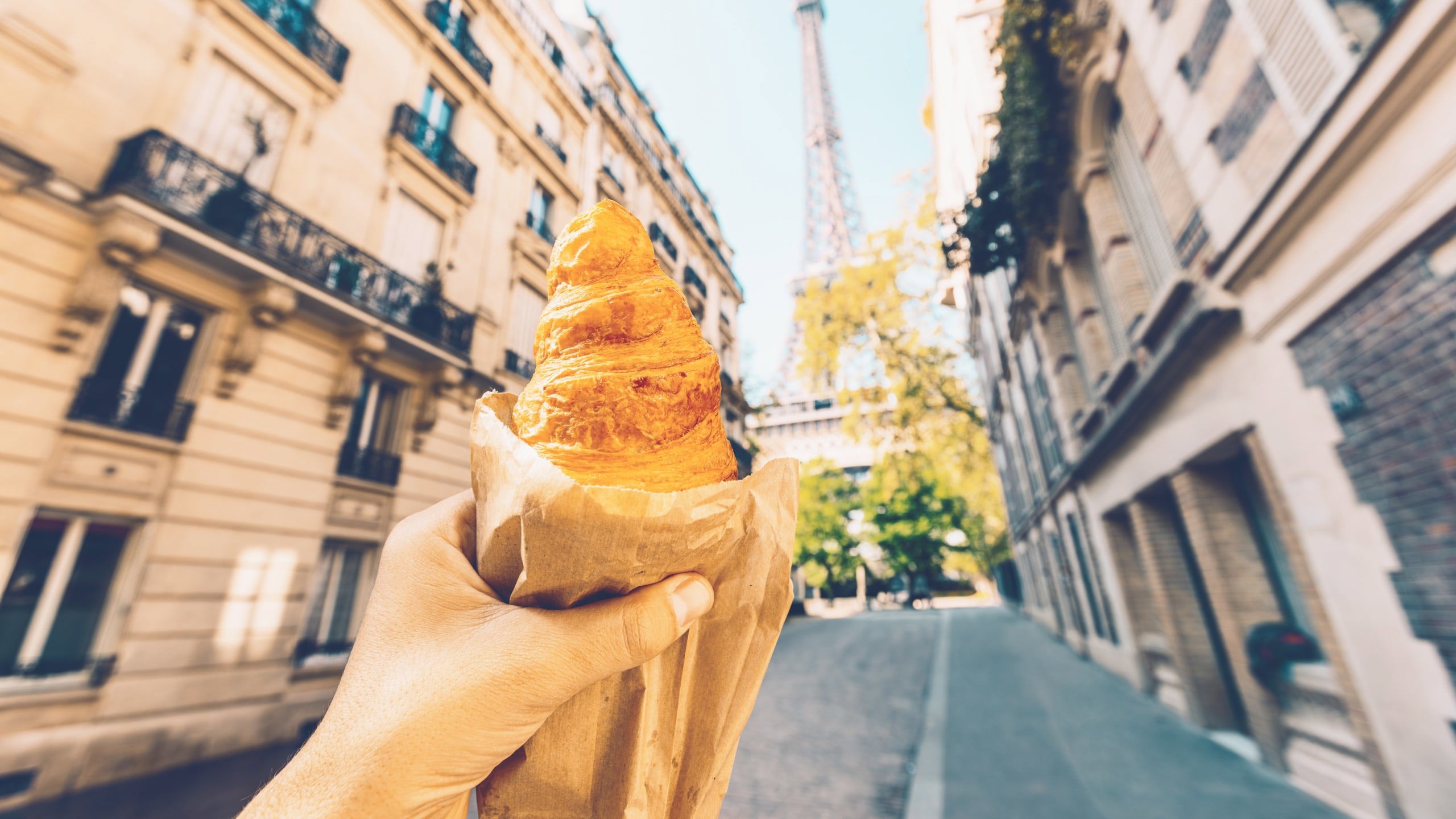 A freshly baked croissant held up towards the Eiffel Tower on a quiet Paris street during the Contiki European Horizon Tour