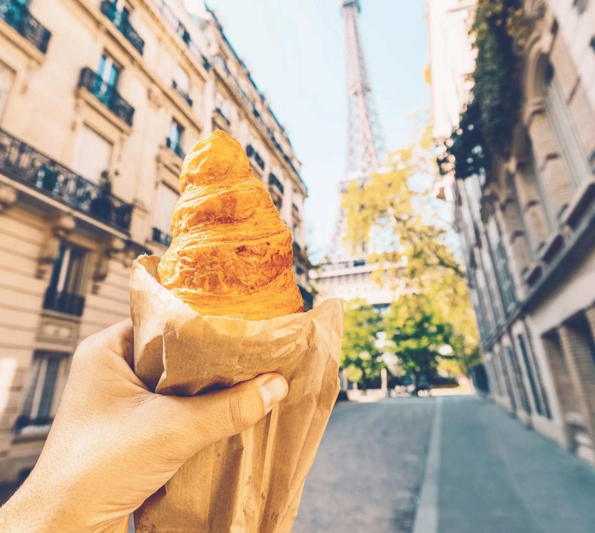 A freshly baked croissant held up towards the Eiffel Tower on a quiet Paris street during the Contiki European Horizon Tour