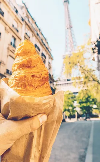 A freshly baked croissant held up towards the Eiffel Tower on a quiet Paris street during the Contiki European Horizon Tour