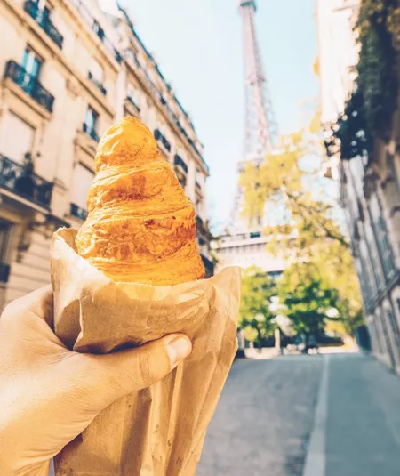 A freshly baked croissant held up towards the Eiffel Tower on a quiet Paris street during the Contiki European Horizon Tour